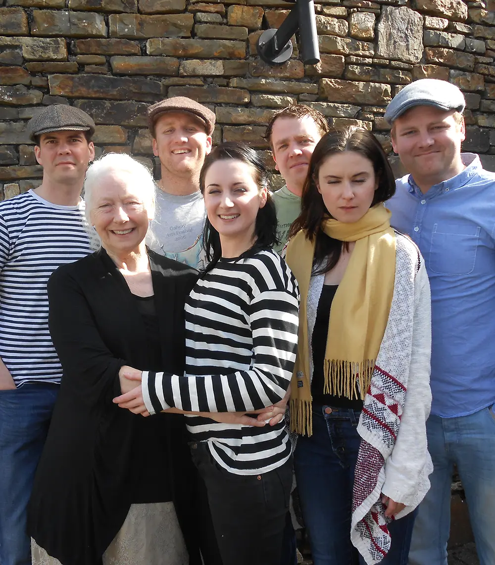 A group of seven people smiling together at the Arts centre Tralee Kerry, near a stone wall.