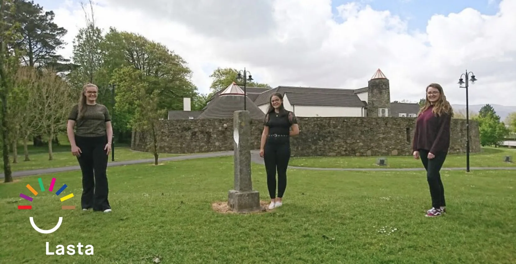 Three women stand on grass near Siamsa Tíre arts centre, with a stone cross and building behind.