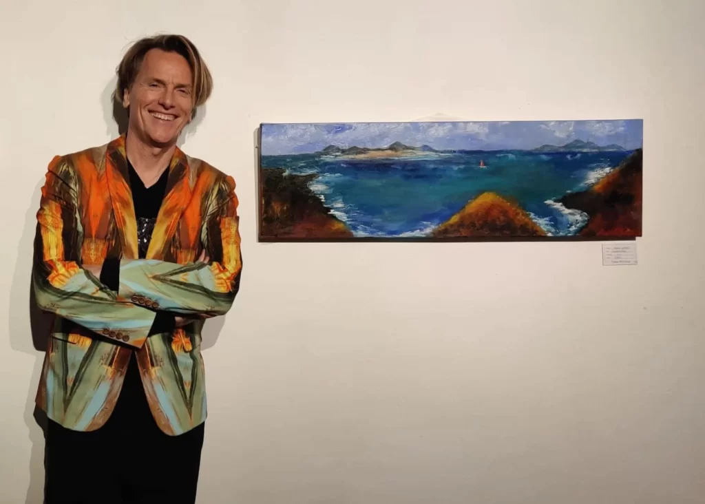 Smiling person in a colorful jacket stands by a seascape painting at Arts centre Tralee Kerry.
