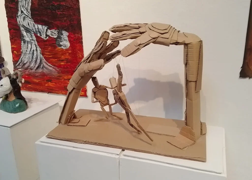 Cardboard hands control a puppet-like figure, echoing Siamsa Tíre traditional Irish arts performances.