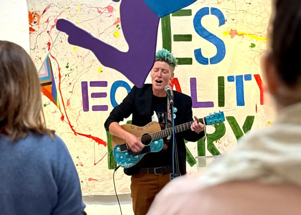 Person with teal hair sings and plays guitar at a vibrant Siamsa Tíre live show.