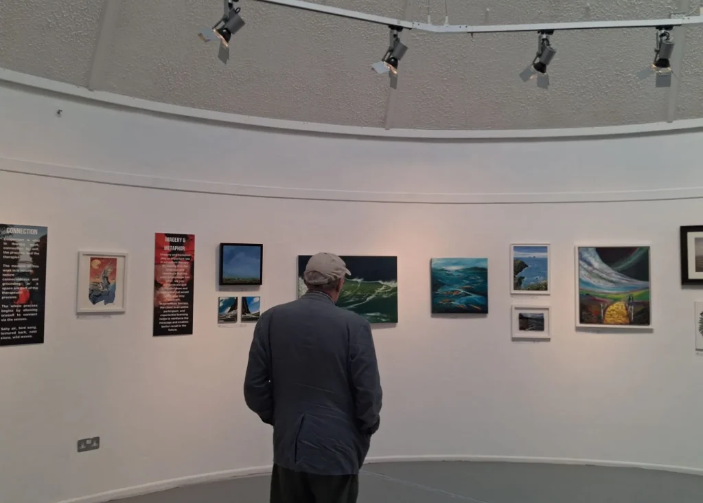 Man wearing a cap views paintings at Siamsa Tíre theatre Tralee in a brightly lit art gallery.