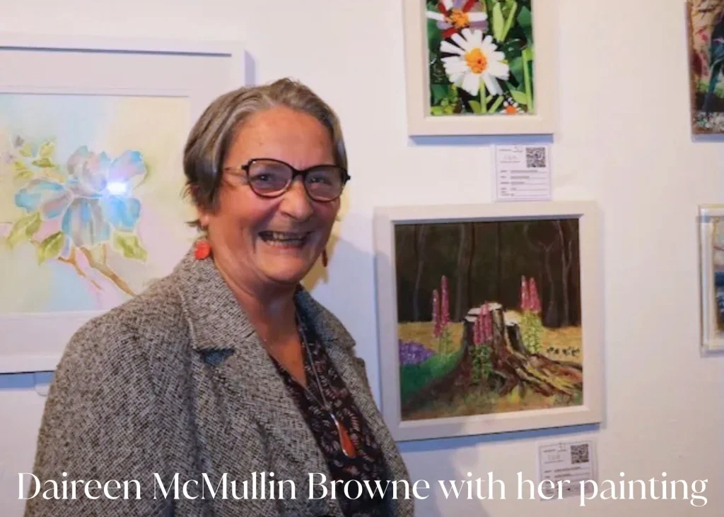 An older lady smiles in front of paintings of flowers hanging on a wall