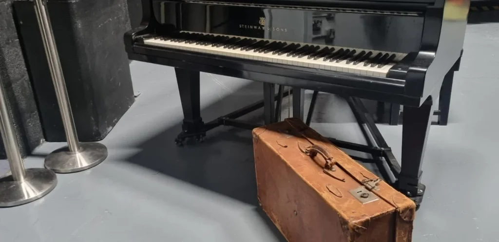 Image of an old brown suitcase beside a black Steinway Piano